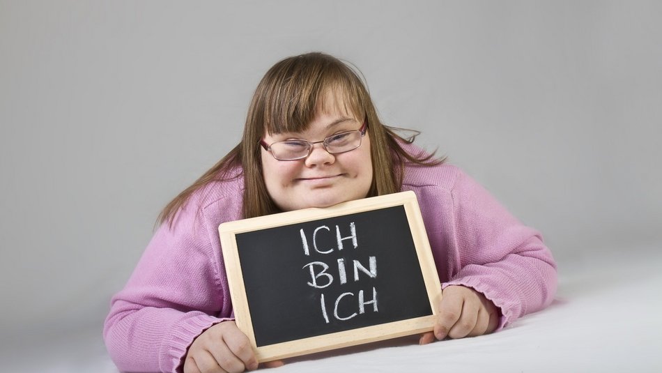 Mädchen mit Downsyndrom liegt auf dem Bauch mit einem Schild in der Hand auf dem Steht: ich bin ich