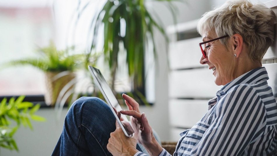 Ältere Frau mit Brille sitz auf dem Boden mit einem Tablet in der Hand